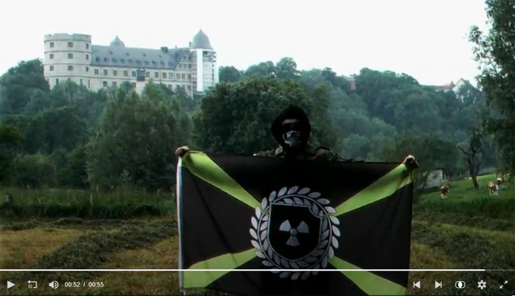 Propaganda-Video von "Atomwaffen Division": Mit Totenkopf-Maske vermummter Anhänger mit Flagge vor der Wewelsburg.