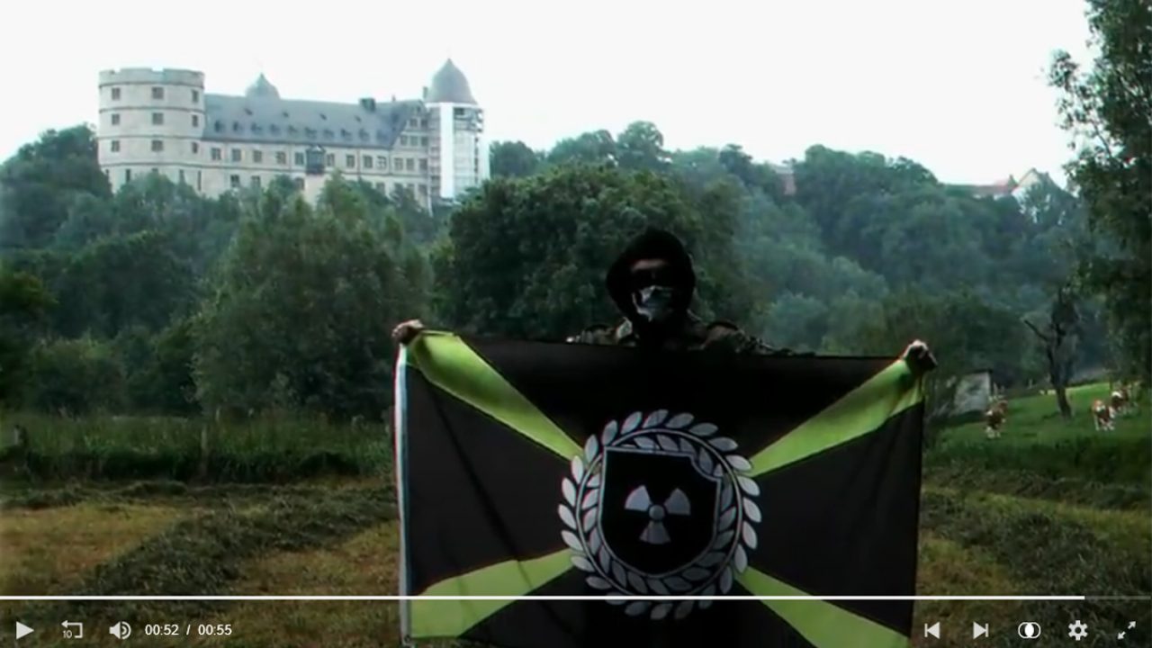 Propaganda-Video von "Atomwaffen Division": Mit Totenkopf-Maske vermummter Anhänger mit Flagge vor der Wewelsburg.