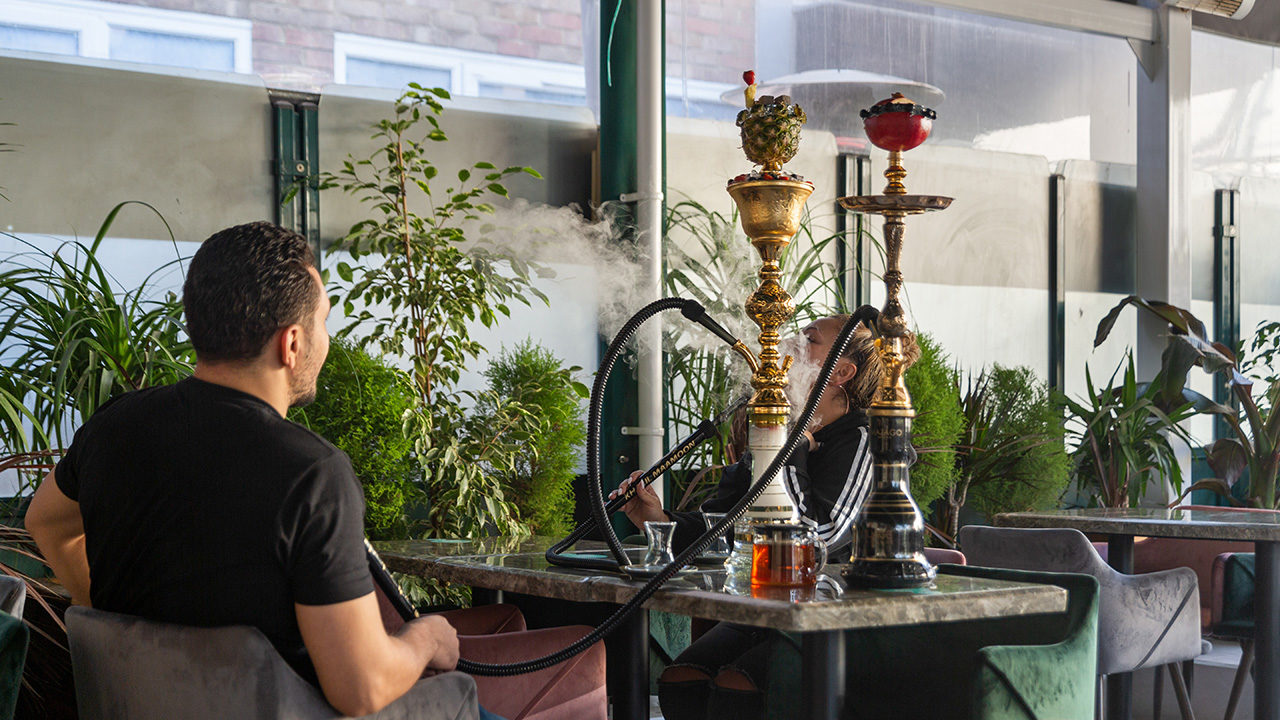 Zwei Menschen sitzen an einem Tisch. Vor ihnen stehen zwei Wasserpfeifen