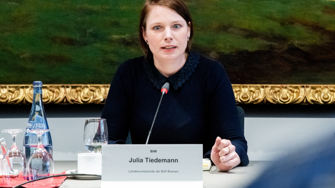 Eine junge Frau sitzt an einem weißen Tisch, vor einem Gemälde mit Goldrand. Sie trägt einen schlichten Pagenkopf und einen schwarzen Pullover und blickt sehr angestrengt. Vor ihr ein Schild mit dem Namen "Julia Tiedemann - BiW".