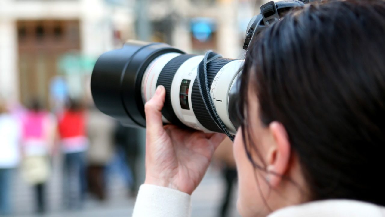 Eine weiblich gelesene Person mit langen Haaren schaut durch den Sucher einer Kamera mit großen Objektiv.